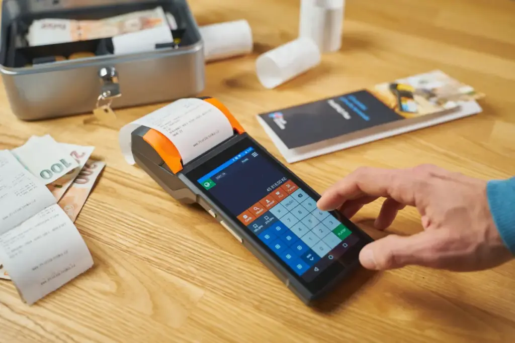 Pessoa usando um terminal de pagamento digital em uma mesa de madeira com dinheiro e recibos visíveis.