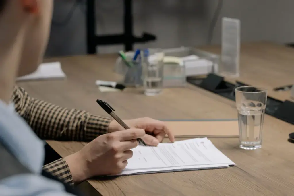 Pessoa vestindo traje social assinando um documento em uma mesa de madeira em um escritório.