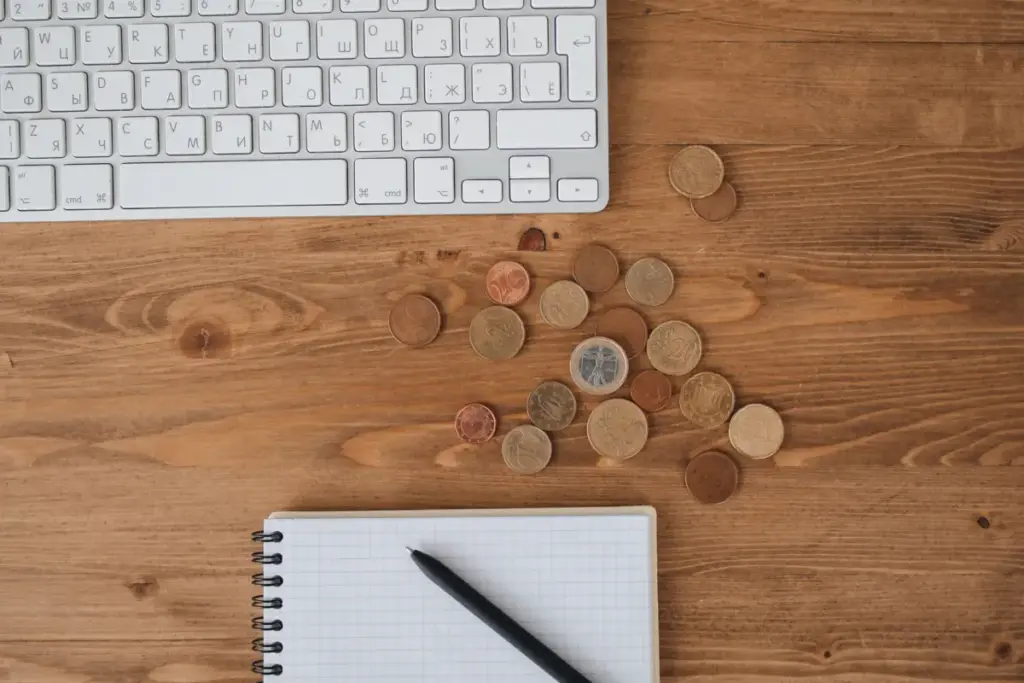 Moedas espalhadas sobre uma mesa de madeira ao lado de um teclado e um caderno, capturados de cima.