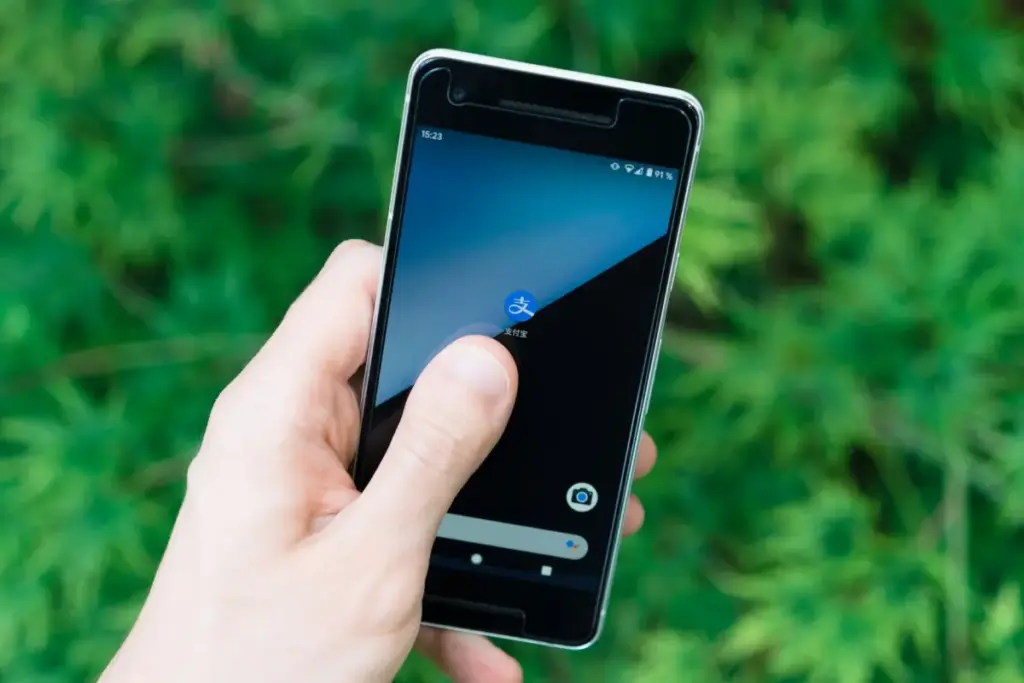 Mão segurando smartphone com ícone de aplicativo na tela, mostrando tecnologia moderna ao ar livre.